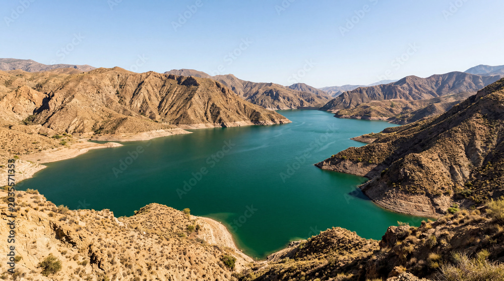 Fototapeta premium Deep Emerald Reservoir Basin Bordered by Sun Baked Clay Hills Under Dramatic Clouds Today