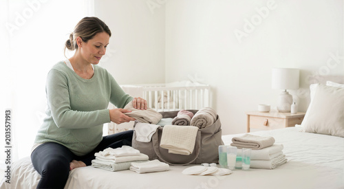Pregnant woman packing hospital bag on bed. Expecting mother preparing baby clothes and toiletries for childbirth in nursery. Maternity and healthcare concept