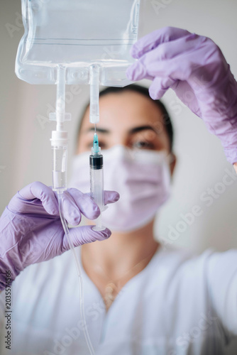 Masked nurse carefully holding syringe near IV bag while preparing intravenous treatment in a bright medical clinic.