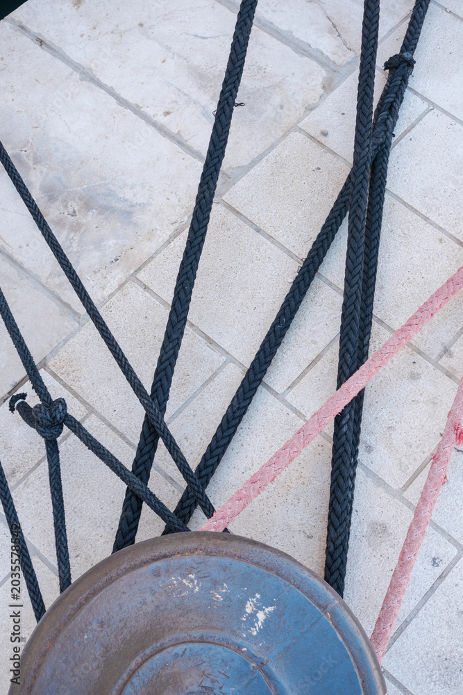 Obraz premium Close up view of several thick mooring ropes tightly tied around an old, rusty bollard on a paved dock