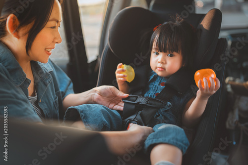 A loving Asian mother carefully buckles her smiling toddler daughter into a secure car seat before a road trip. Concept of child safety, parenting, and family travel security