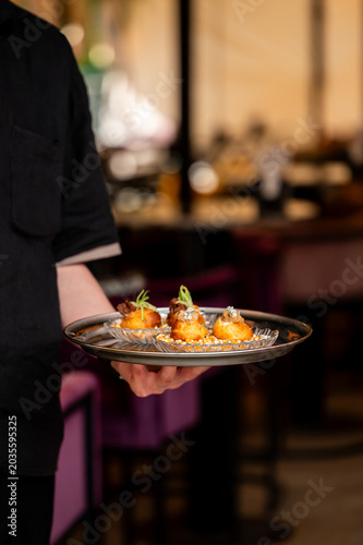 A waiter in a black shirt holds a silver tray with gourmet appetizers served on small glass plates in a restaurant.