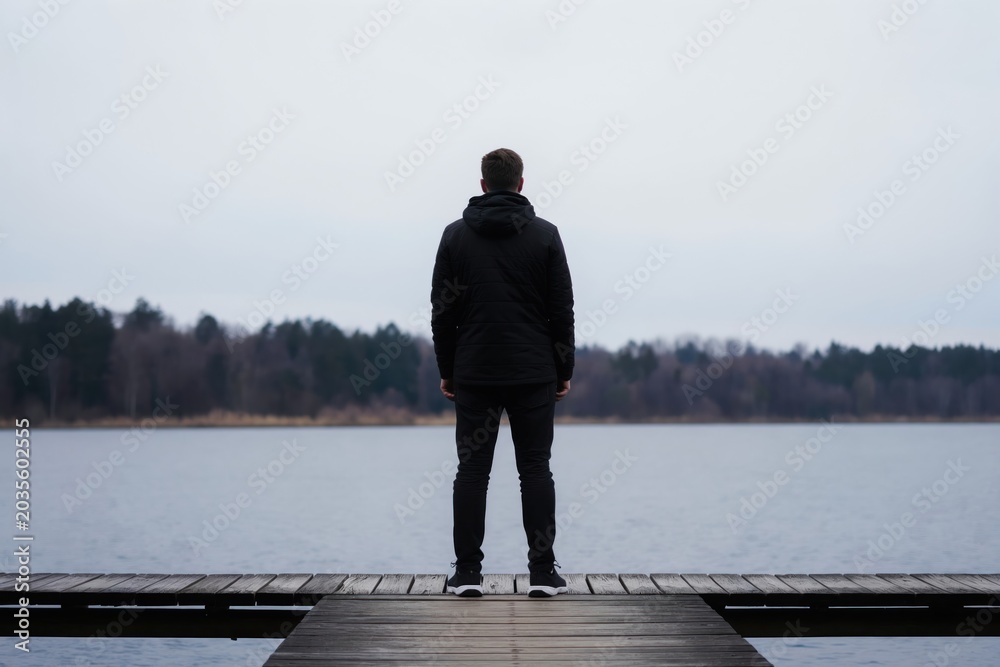 Fototapeta premium Man in black jacket stands on wooden pier over lake. He looks at water with thoughtful expression. Peaceful winter nature scene with forest backdrop. Solitary figure, quiet contemplation, fresh air.