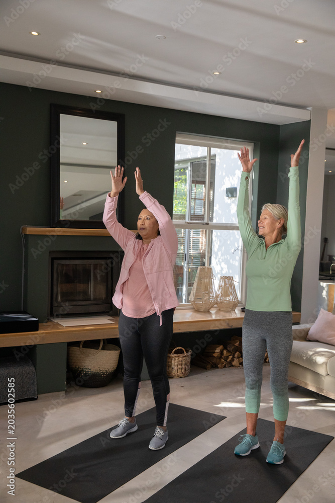 Fototapeta premium Female friends standing on black yoga mats in living room stretching overhead near fireplace mantel