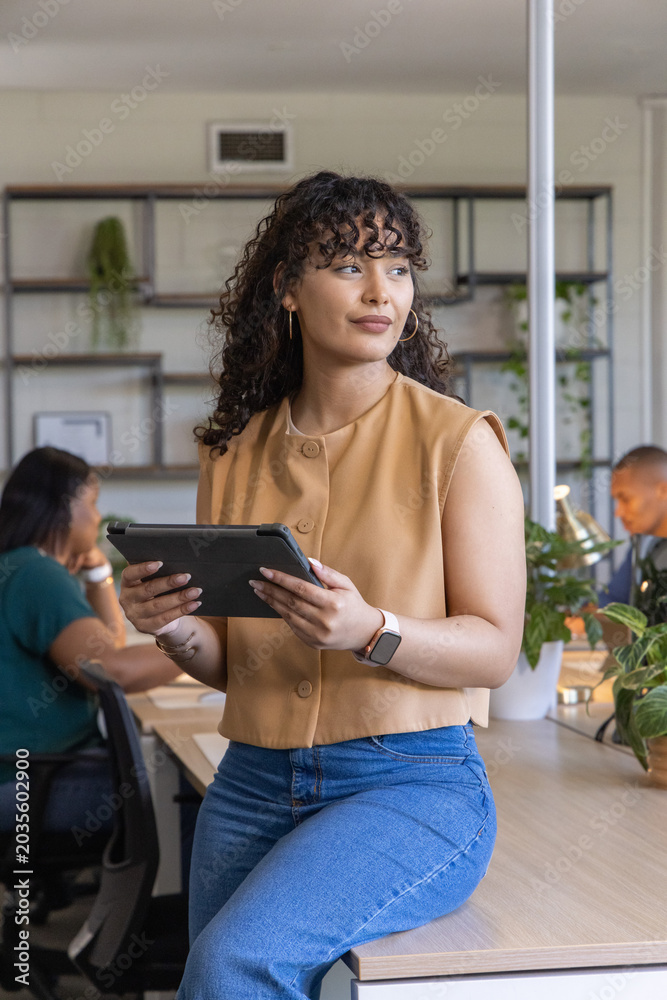 Fototapeta premium Diverse coworkers with Hispanic woman sitting on desk edge holding tablet in open-plan office