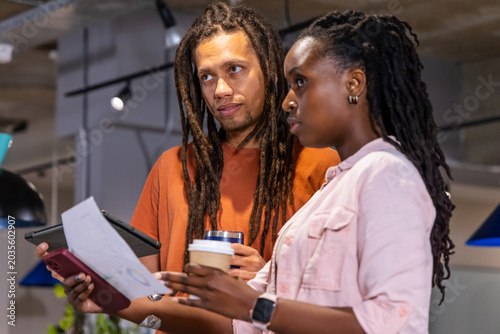 Diverse coworkers reviewing printed document and tablet while holding smartphone and coffee cup