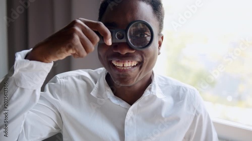 Cheerful black man smiles in an office, wearing a white shirt. He playfully holds a magnifying glass over one eye, humorously enlarging it while looking directly at the camera.