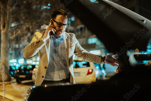 Schilderij op canvas Businessman making a phone call, standing next to a broken down car with its hoo