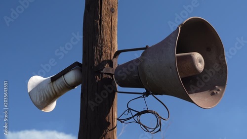 Vintage aluminum loudspeaker attached to a wooden utility pole against blue sky. Slow camera movement from left to right. Retro public announcement and communication concept.