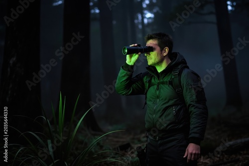 Man with night vision device searches dark forest at night. Guy uses optical monocular for spotting wildlife or intruders in woods. Tech equipment helps see in low light conditions.