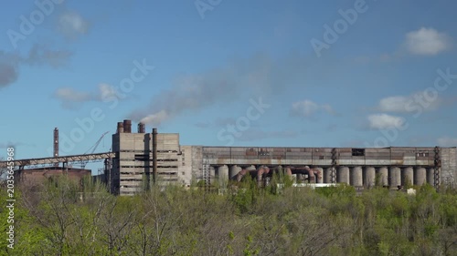 Static shot of industrial factory complex with smokestacks and industrial buildings surrounded by green trees under blue sky. Heavy industry landscape footage in 4K.