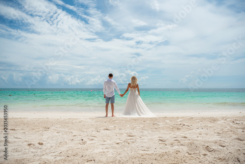 Newlyweds standing on white sand beach facing turquoise sea