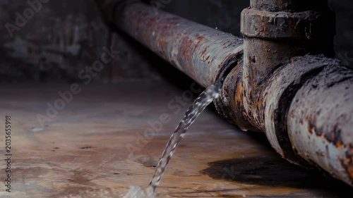 A rusted industrial pipe leaks water onto a dry concrete floor inside a building. The flow creates small puddles and runs across the surface, highlighting the decay and wear of the pipe