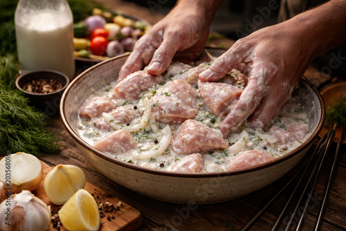 Hands marinating raw chicken pieces in creamy herb marinade with onions in ceramic bowl on rustic wooden table with fresh ingredients.