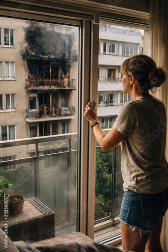 Woman looking at fire damaged apartment building from balcony window holding coffee mug in urban residential area.