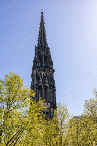 The towering blackened spire of St. Nikolai Memorial rises above green foliage under a clear blue sky in Hamburg, Germany.
