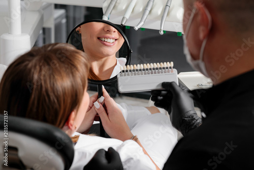 a smiling woman at a dental clinic holds a mirror while a dentist in black gloves shows her a tooth shade guide