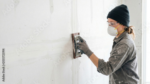 Woman sanding drywall wall with sander tool, focused on finishing touches to smooth surface. Drywall installation includes preparing walls for painting and decoration