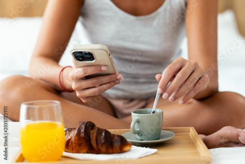 Woman using a smartphone while having breakfast on bed at home.