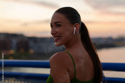 Slika na platnu Smiling woman in blue sports bra and wireless earbuds standing on bridge