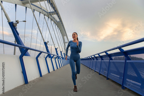 Foto Young woman jogging on modern pedestrian bridge at sunset, wearing blue activewe