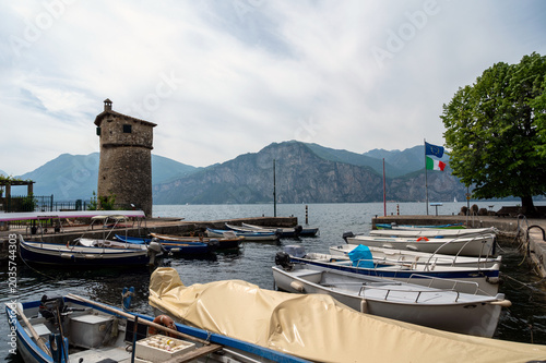 Small boats moored at a Lake Garda harbor in Italy with a historic stone tower, Italian flags, and mountain view.