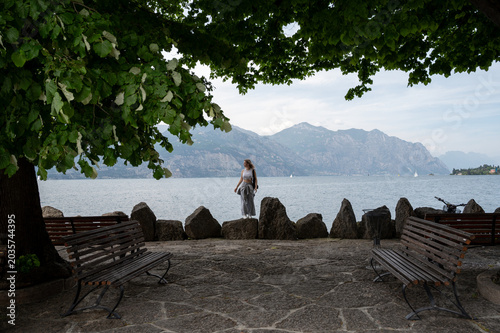 Woman standing by a lakeside viewpoint on Lake Garda, Italy, with benches, trees, mountains, and calm water in the background.
