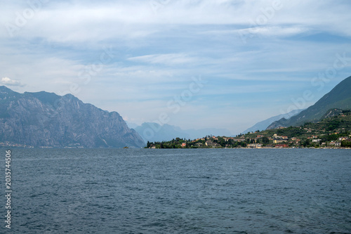 Wide panorama of Lake Garda in Italy with a lakeside town, mountains, and calm water under a bright summer sky.