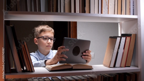 Photos Young boy in glasses focusing on a digital tablet screen, sitting at a bookshelf