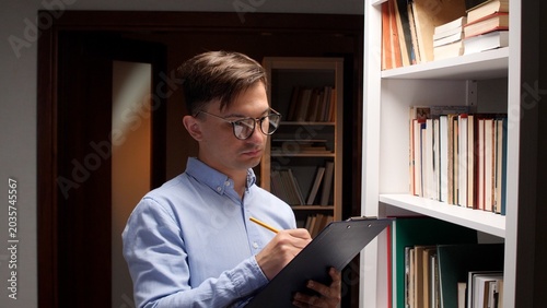 Man wearing glasses intently focusing, writing notes with a pencil on a clipboard while standing by a bookshelf filled with books in a library, conducting inventory or research