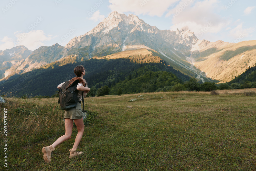 Naklejka premium Woman hiking through a grassy meadow toward rugged mountains, carrying a backpack, enjoying fresh air and wide landscapes in a nature adventure scene.