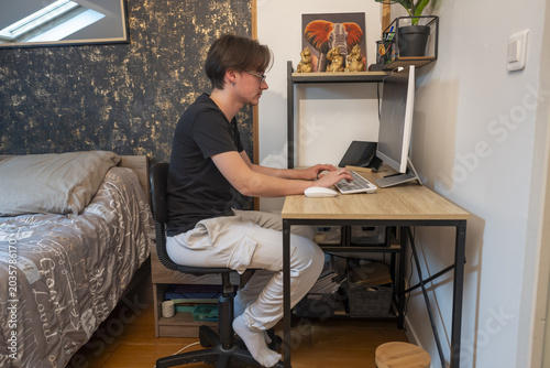 Teenage boy sitting at a desk using a laptop in his bedroom, studying or working from home in a casual interior.