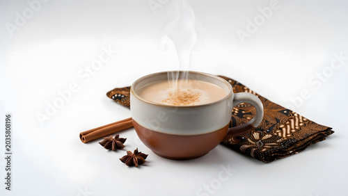 Steaming cup of chai tea with cinnamon stick and star anise on a patterned cloth, against a white background.