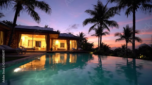Eco luxury tropical villa architecture with illuminated pool, palm trees, and glowing sunset reflecting calm water evening light