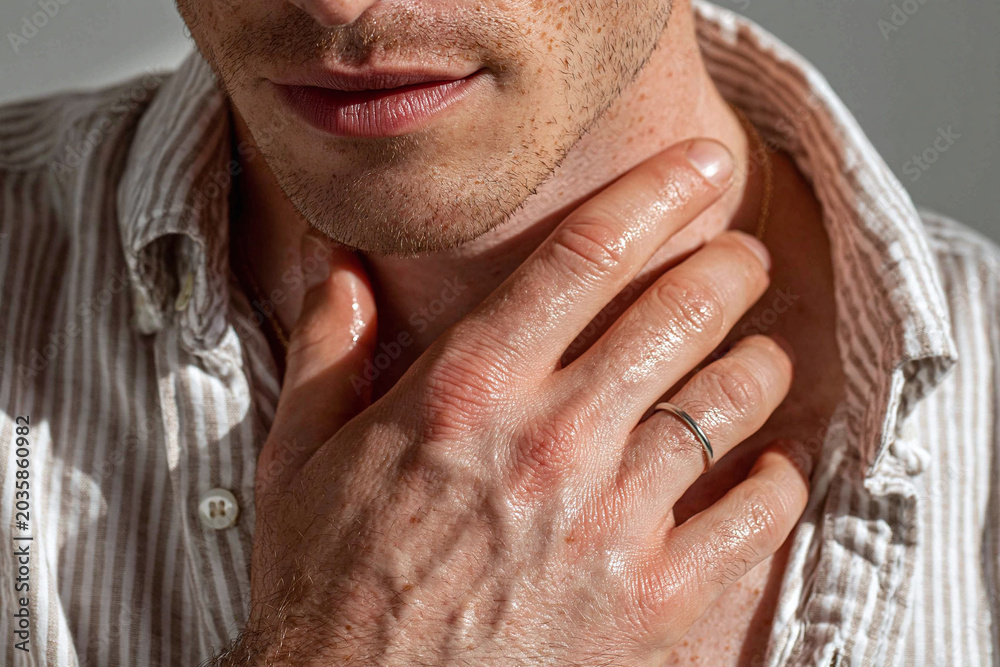Fototapeta premium Man hand placed on collarbone sunlight grazing skin, close up of male chest and lips, textured stubble and ring