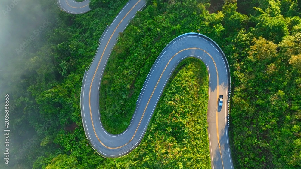 Obraz premium Aerial drone view of a stunning winding mountain road through a misty tropical forest at sunrise, with a car climbing the steep path, symbolizing transportation, and adventure. Nan Province, Thailand.