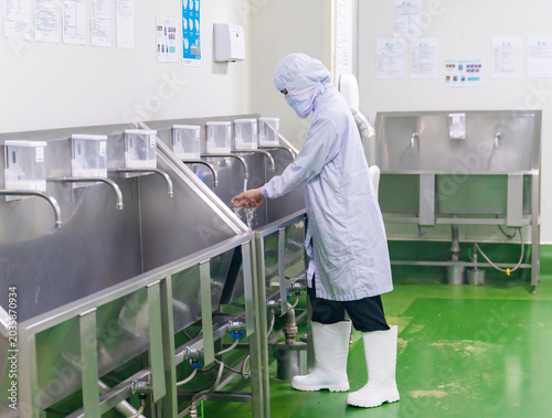 A food processing plant worker in full protective uniform, hairnet, mask, and white rubber boots washes their hands at a stainless steel industrial sink station before entering the production line.