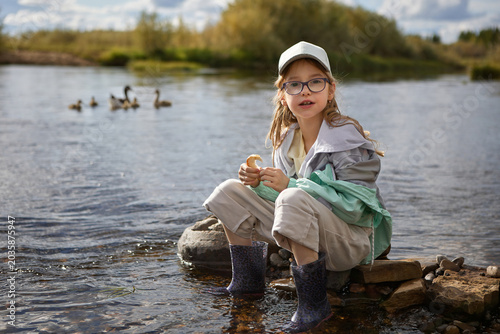 Fotografie Little girl with glasses sitting by a river in rubber boots and casual outdoor clothes