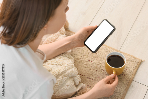 Young woman with yellow cup of coffee and mobile phone lying on carpet at home