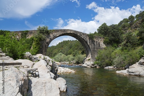Steinbrücke über einem ruhigen Fluss mit felsiger Uferlandschaft und dichtem Grün unter einem blau bewölkten Himmel, Römische Brücke, Piscina natural de Madrigal de la Vera, Naturbad Madrigal de la Vera, Garganta de Alardos, Schlucht Alardos, Cáceres, Extremadura, Spanien