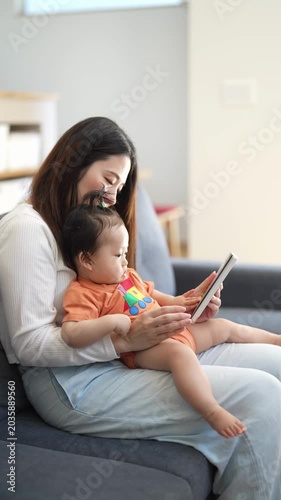 Mother smiles warmly while holding child. Child gazes at tablet with quiet curiosity. They sit close on soft couch, sharing quiet moment. Perfect for parenting, family, or tech use