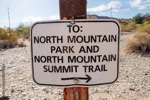 Closeup view of a weathered metal trail sign directing hikers to North Mountain Summit, standing against rocky desert terrain, sparse vegetation, and distant hills beneath soft clouds. Phoenix, Arizon