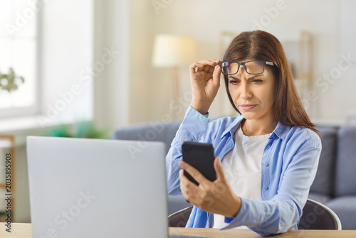 Принти на полотні Woman with myopia, presbyopia and vision loss holding glasses on forehead to peer at and read text on mobile phone screen, struggle with blur and eyestrain