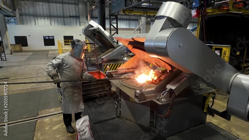 Cinematic industrial footage featuring molten metal pouring from a high-temperature furnace inside a modern manufacturing facility. Intense heat, glowing steel, sparks, heavy machinery, and industrial