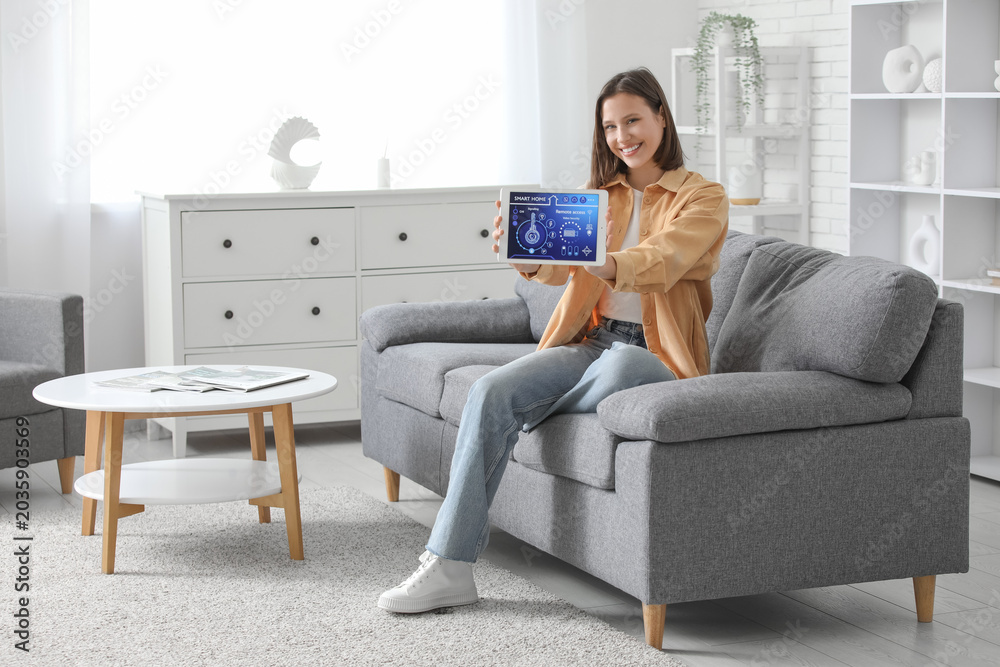 Obraz premium Young happy woman holding tablet with smart home security system control panel on sofa in room