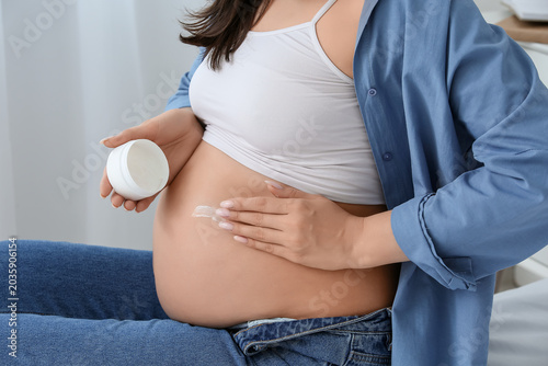 Young pregnant woman applying cosmetic cream in bedroom, closeup