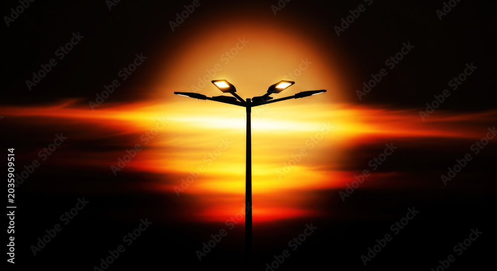 Fototapeta premium A tall street light stands silhouetted against a vibrant, fiery sunset. Two light fixtures illuminate the darkening sky, casting a warm glow.