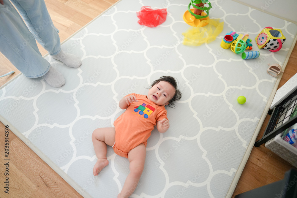 Naklejka premium Baby lies smiling on soft patterned mat. Tiny hands reach toward colorful toys nearby. Adult feet stand close, watching calmly. Bright yellow ball and plush items scattered around