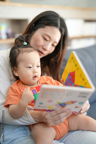 Mother holds baby, reading colorful book together. Baby looks up with wonder, fingers touching pages. Soft light, cozy living room, gentle moment shared. Perfect for parenting, education