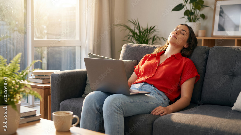 Obraz premium Young woman resting on a sofa with a laptop in a sunlit living room